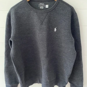 Mörkgrå sweatshirt från Polo Ralph Lauren - Mörkgrå sweatshirt från Polo Ralph Lauren med klassisk rund hals och broderad vit logga på bröstet. Tröjan har lång ärm och är tillverkad i mjuk bomullsblandning, perfekt för en chill och stilren look.