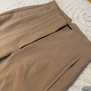 Beige kostymbyxor från Zara - Snygga beige kostymbyxor från Zara med hög midja och raka ben. Byxorna har diskreta fickor fram och stängs med dragkedja, hake och knapp. Perfekta för en clean och stilren look. Materialet känns mjukt och har en lätt stretch.