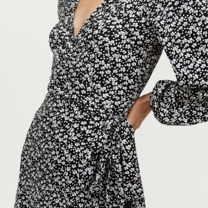 Blommig omlottklänning  - Supersöt vintage omlottklänning från Gina Tricot i marinblått med vita småblommor. Klänningen har långa ärmar med volangdetalj vid ärmslut, knyt i midjan och v-ringning. Längd: 110 Midja: 100 cm OBS! Första bilden är inte samma klänning, bilden visar bara hur passformen på klänningen ser ut. 