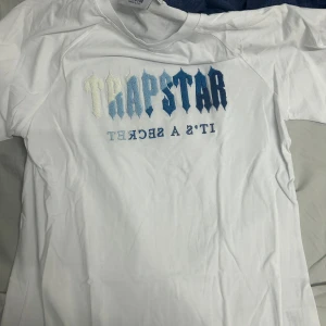 Vit Trapstar t-shirt med blått tryck - Snygg vit t-shirt från Trapstar med stort broderat logotyp-tryck i blå och vita nyanser på bröstet. Klassisk passform och korta ärmar, perfekt för en streetwear-look. Materialet känns mjukt och skönt mot huden.