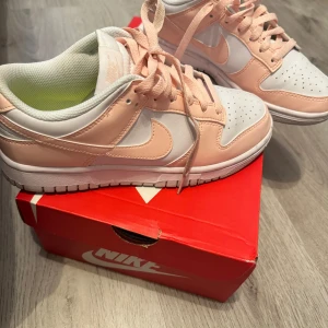 Nike Dunk Low i vitt och ljusrosa - Nike Dunk Low sneakers i en fräsch kombo av vitt och ljusrosa skinn. Klassisk siluett med rund tå, snörning och vit sula. Insidan har en limegrön innersula med svart Nike-logga. Perfekta för dig som vill sticka ut med stilrena färger. Använd en gång