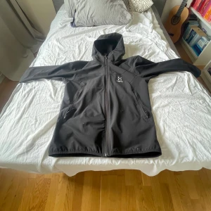 Svart vindjacka från Haglöfs, stl L - Svart vindjacka från Haglöfs i storlek L. Jackan har huva, hel dragkedja framtill och två dragkedjeförsedda fickor. Tillverkad i slitstark polyester och perfekt för blåsiga dagar. Snygg minimalistisk design med Haglöfs-logga på bröstet. Den är L men passar S för det är en unisex jacka så då skriver dom L men den passar S och m