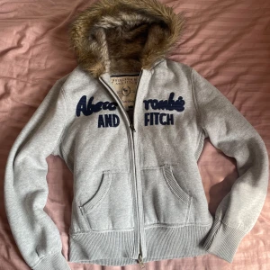 Grå hoodiejacka Abercrombie & Fitch XL - Grå hoodiejacka från Abercrombie & Fitch med dragkedja, två fickor och en huva fodrad med mjuk fuskpäls. storleken är xl men den passar på mig som brukar ha s i tröjor. skriv vid frågor och lägg gärna prisförslag!