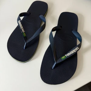 Mörkblå flip-flops från Havaianas - Snygga mörkblå flip-flops från Havaianas med klassisk design och brasiliansk flagga på remmen. Tillverkade i syntetmaterial med bekväm platt sula och diskret mönstrad yta. Perfekta för sommardagar och strandhäng.
