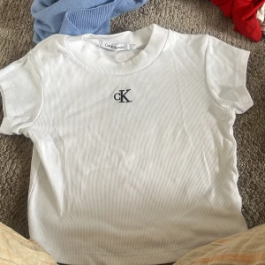 Vit ribbad t-shirt från Calvin Klein - Vit ribbad t-shirt från Calvin Klein Jeans med broderad svart cK-logga på bröstet. Klassisk rund halsringning och korta ärmar. Snygg och enkel modell som är lätt att matcha till allt.