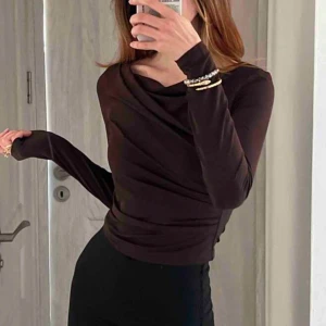 Brun långärmad topp med drapering - Snygg brun långärmad topp med draperad effekt över bröstet och axlarna. Tunn och stretchig, sitter tight och har en elegant, asymmetrisk halsringning. Perfekt att styla med både jeans och kostymbyxor för en trendig look.