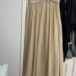 Champagnefärgad långklänning med pärlor - Elegant långklänning i champagnefärg med vackra pärldetaljer upptill. Klänningen har en romantisk och flowy siluett, perfekt för bal eller fest. Axelbandslös design som ger en stilren och lyxig känsla.
