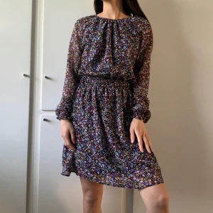 Blommig kortklänning med resårmidja - Supersöt kortklänning med småblommigt mönster i lila, gult och grönt på mörk botten. Långärmad modell med resår i midjan och vid ärmslut. Tunnt tyg, med underkjol. Har en knapp i nacken.