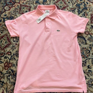 Rosa pikétröja från Lacoste - Snygg rosa pikétröja från Lacoste med klassisk krage och två knappar framtill. Ikoniska krokodilloggan broderad på bröstet. Tröjan är i bomull och har korta ärmar, perfekt för en fräsch och sportig look