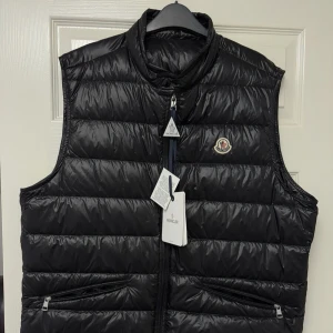 Moncler Gui - Snygg svart dunväst från Moncler med hög krage och två dragkedjefickor framtill. Västen har quiltad design och klassisk Moncler-logga på bröstet. Perfekt för lager-på-lager och ger en clean look till din outfit.