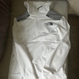 Vit hoodie från The North Face M - Snygg vit hoodie från The North Face med grå axelpartier och logga på bröstet. Tröjan har huva, lång ärm och half zip-dragkedja framtill. Perfekt för dig som gillar sportig och clean stil. Storlek M