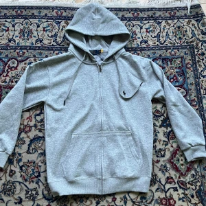 Grå hoodie från Polo Ralph Lauren - Snygg grå hoodie från Polo Ralph Lauren med dragkedja och klassisk huva. Tröjan har snörning i huvan, två stora fickor framtill och den ikoniska broderade loggan på bröstet. Perfekt för chill dagar och enkel att matcha med jeans eller joggers.