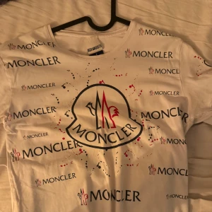Vit Moncler t-shirt med logga - Snygg vit t-shirt från Moncler med stort tryck av loggan och flera mindre Moncler-tryck över hela tröjan. T-shirten har korta ärmar och rund halsringning. Färgdetaljer i svart och rött ger en cool kontrast till den vita basen.