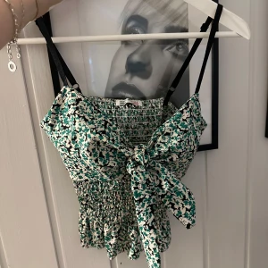 Blommig grön och vit linnetopp med rosett - Supersöt linnetopp med grönt och vitt blommönster, smockad midja och stor rosett framtill. Smala svarta axelband och volangkant nedtill ger en extra gullig touch. Perfekt för varma dagar och snygg att styla med jeans eller kjol.