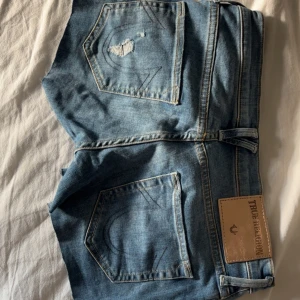 Blå jeansshorts från True Religion - Snygga blå jeansshorts från True Religion med klassiska bakfickor och slitna detaljer. Shortsen har låg midja, råa kanter och en normal passform. Perfekta för sommaren. Egentligen skinny jeans men gjorde om de till shorts. Kom privat för mer bilder. 