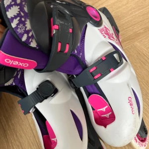 Oxelo Play 5 inlines i vitt och lila - Snygga inlines från Oxelo i vitt, lila och rosa med blommigt mönster och svarta detaljer. Skorna har justerbara spännen och robust plastskal för extra stöd. Perfekta för dig som vill rulla fram med stil och färg.