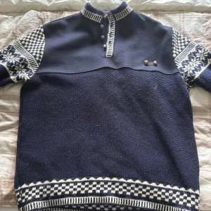 Mörkblå norsk stickad tröja Score XL - Snygg mörkblå stickad tröja från Scandinavian Score Design med vita mönster på ärmar och nederkant. Tröjan har hög krage med knappar och klassiska norska detaljer. Perfekt för kalla dagar och har en autentisk badge på ärmen. Tröjan är använd ett fåtal gånger. Det är bara att skriva om ni undrar något. Pris kan diskuteras vid en snabb affär 