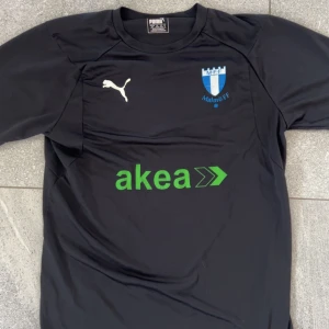 Svart Malmö FF fotbollströja Puma - Svart fotbollströja från Puma med Malmö FF-logga på bröstet och sponsortryck 'akea' i grönt. Tröjan har korta ärmar och är tillverkad i ett lätt, tekniskt material som andas. Perfekt för träning eller match.
