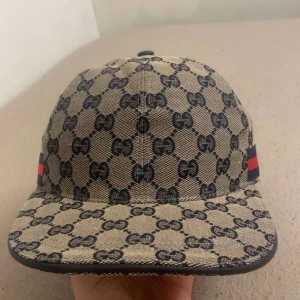 Gucci keps med GG-mönster och röd-blå detalj - Snygg keps från Gucci i beige och marinblått med klassiskt GG-monogram över hela kepsen. Kepsen har en röd och marinblå rand på sidan och justerbar rem baktill i skinn. Tillverkad i bomullscanvas med innerfoder i marinblått tyg. Perfekt för dig som vill sticka ut med lyxig streetstyle.