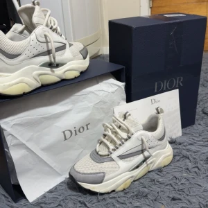 Dior B22 - Snygga chunky sneakers från Dior i vitt och grått med mesh och skinnpaneler. Skorna har en kraftig sula, rund tå och coola detaljer som snörning och logga på sidan. Perfekta för dig som gillar statement-sneakers med lyxig känsla. Storlek 42,5 hör av er! Så löser vi en bra deal😁
