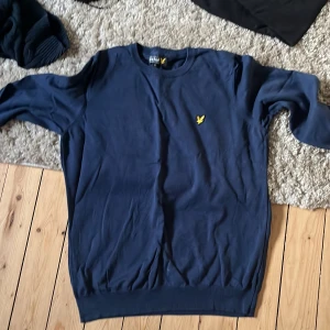 Lyle & Scott tröjor i blått och svart XL - Två klassiska crewneck-tröjor från Lyle & Scott i storlek XL. En är marinblå och en är svart, båda med den ikoniska gula örnloggan på bröstet. Tröjorna är i mjuk bomull och har ribbade muddar vid ärmslut och nederkant. Perfekt för en clean och stilren look.
