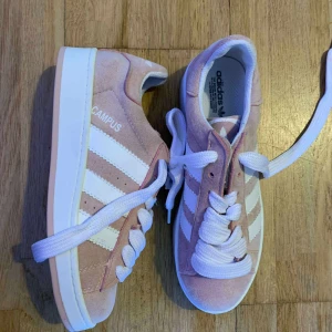 Adidas Campus rosa sneakers - Snygga Adidas Campus sneakers i ljusrosa mocka med klassiska vita ränder och vit sula. Skorna har rund tå och breda vita snören för en cool look. Perfekta för dig som vill ha en stilren och trendig sneaker med retrovibbar.