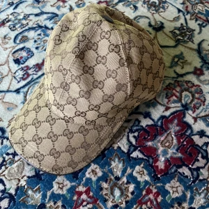 Beige keps från Gucci med GG-mönster - Snygg beige keps från Gucci med det klassiska GG-monogrammet över hela kepsen. Kepsen har böjd skärm och är tillverkad i ett slitstarkt bomullsmaterial. Perfekt för dig som vill ha en exklusiv och trendig accessoar med tydlig logga.