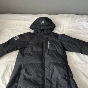 Svart dunjacka från Sail Racing M - Svart dunjacka från Sail Racing med huva och hel dragkedja. Jackan har en clean look med logga på bröstet och ärmen, samt quiltad design. Perfekt för kalla dagar och riktigt snygg till streetwear-stilen.