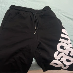 Svarta shorts från Dsquared2 med tryck - Svarta shorts från Dsquared2 med stor vit logotyp längs ena benet. Modellen har elastisk midja med snörning och är tillverkad i mjukt material, perfekt för en avslappnad och sportig stil. Snygga och bekväma shorts med fickor på sidorna.