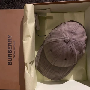 Beige rutig keps från Burberry - Snygg beige keps från Burberry med diskret rutigt mönster och broderad logga baktill. Klassisk böjd skärm och justerbar rem bak. Tillverkad i bomull för en bekväm och stilren look. Perfekt accessoar för att lyfta din outfit.