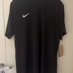 Svart Nike Dri-FIT t-shirt XL - Nike Dri-FIT Träningtshirt – Svart En stilren och funktionell träningströja från Nike i klassisk svart färg med vitt swoosh-logotyptryck på bröstet. Tillverkad i lätt, snabbtorkande Dri-FIT-material som effektivt transporterar bort svett och håller dig torr under hela träningspasset. Perfekt för gym, löpning, fotboll eller annan fysisk aktivitet. ✔ Normal passform ✔ Andasmaterial ✔ Dri-FIT-teknologi ✔ Passar både inomhus- och utomhusträning ✔ Diskret och stilren design utan tryck på ryggen 