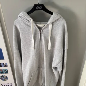 Grå hoodie med dragkedja från H&M - Säljer en ljusgrå hoodie från H&M i storlek S. Tröjan har huva med vita snören, dragkedja framtill och två fickor. Perfekt för chill dagar eller när du vill ha en enkel och clean look. Materialet är mjukt och skönt, troligtvis bomullsmix.