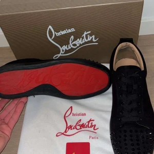Christian Louboutin svarta sneakers med nitar - Svarta sneakers från Christian Louboutin i mocka med ikonisk röd sula. Skorna har svarta nitar på tån, svarta skosnören och en rund tå. Insidan är beige och designen är riktigt edgy och lyxig – perfekt för dig som vill sticka ut. Är ni intresserade är det bara att skriva till labenclozet
