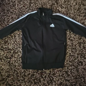 Svart Adidas med vita ränder - Svart tröja från Adidas med klassiska vita ränder längs ärmarna och broderad logga på bröstet. Jackan har dragkedja hela vägen, ståkrage och två fickor med dragkedja. Perfekt för en sportig look.