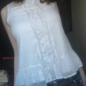 Vit ärmlös blus - Supersöt vit blus i transparent tyg med små prickar, volang nertill och spets längs knäppningen framtill. 