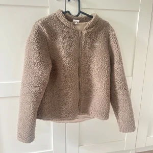 Brun teddyjacka - Mysig brun teddyjacka från Aimn med dragkedja framtill och rundad krage. Jackan är fluffig och har en enkel, stilren look utan synliga fickor. Perfekt för dig som vill ha en varm och trendig jacka 