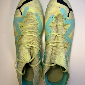Puma Future FG/AG - Säljer ett par snygga Puma fotbollsskor i storlek 39. Skorna är ljusgula med turkosa detaljer och svarta Puma-loggor. De har snörning, platt sula med dobbar och en stickad ovandel för skön passform. Perfekta för dig som vill sticka ut på planen!