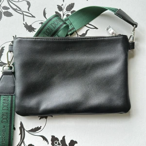 Svart axelväska från Don Donna - Stilren svart axelväska från Don Donna med bred grön axelrem med logga. Väskan är i slätt skinn och har dragkedja upptill samt innerfack för småprylar. Perfekt storlek för mobil, plånbok och nycklar. Snygg och enkel design som funkar till många outfits.