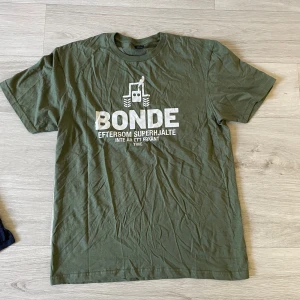 grön t-shirt med vitt tryck på bröstet där det står ett bonde citat - Säljer en grön t-shirt med vitt tryck på bröstet där det står 'BONDE eftersom superhjälte inte är ett erkänt yrke' och en traktor. Klassisk passform och rund halsringning, perfekt för dig som gillar humor och lantliv.