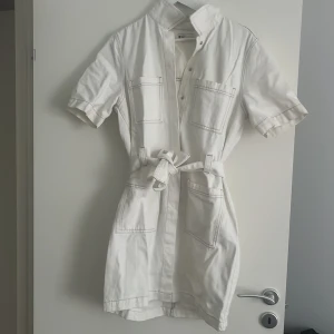 Vit kort skjortklänning från gina tricot - Snygg vit kort skjortklänning från gina tricot med knappar framtill, stora fickor och knytskärp i midjan. Klänningen har korta ärmar och markerade sömmar som ger en cool och modern look. Perfekt för dig som gillar stilrena och trendiga plagg.