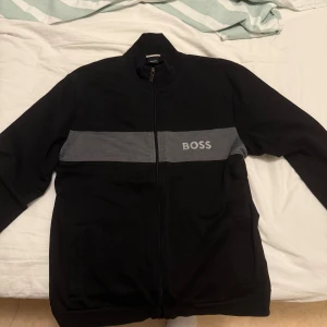 Svart jacka med grå detalj från BOSS - Svart tröja från BOSS med en bred grå rand över bröstet och logga. Tröjan har hel dragkedja, hög krage och två fickor framtill. Tillverkad i mjukt material, perfekt för en clean och stilren look. Köpt på NYL Man 