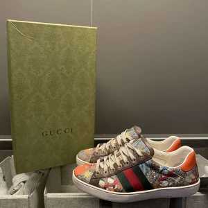 Gucci sneakers storlek 41,5 - Unika Gucci sneakers med färgglatt tecknat mönster, klassiska gröna och röda ränder på sidan och orange detalj vid hälen. Skorna har vita snören och vit sula. Perfekt för dig som vill sticka ut med lyxig streetstyle. Allt og tillkommer dock inget kvitto kvar men box och snören ingår i köpet.