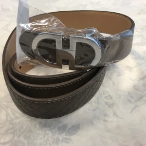 Brunt skärp med silverfärgat spänne - Snyggt brunt bälte med ett stort, silverfärgat spänne i metall. Bältet har ett diskret rutmönster och är gjort i skinn. Insidan är ljusbeige. Perfekt accessoar för att lyfta din outfit. Helt ny 