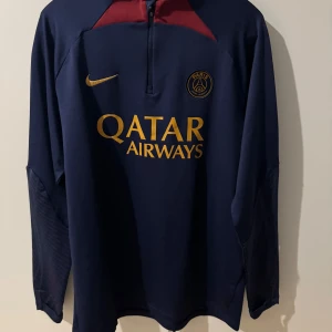 Paris Saint-Germain långärmad träningströja - Mörkblå långärmad PSG träningströja från Nike med gul logga och Qatar Airways sponsortryck. Tröjan har dragkedja vid halsen, röda detaljer upptill och är tillverkad i Dri-FIT material för bra ventilation.