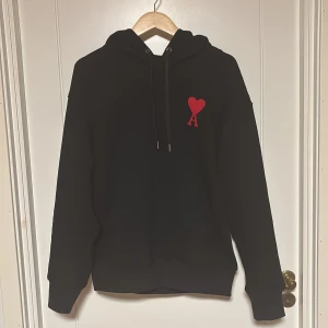 Svart hoodie från Ami Paris - Säljer en svart hoodie från Ami Paris med röd broderad logga i form av ett hjärta och bokstaven A på bröstet. Tröjan har huva med dragsko och klassisk passform. Perfekt för en stilren och avslappnad look.