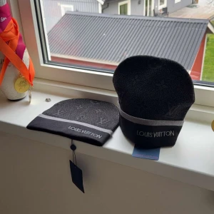 Svart mössa från Louis Vuitton - Snygg svart mössa från Louis Vuitton med diskret monogrammönster och en grå rand med logotypen broderad framtill. Tillverkad i mjukt material som känns lyxigt och bekvämt. Perfekt accessoar för att lyfta din stil.