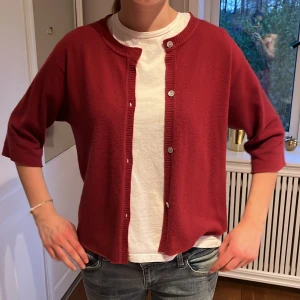 Mörkröd kofta med knappar - Säljer en mörkröd kofta med rund hals och knappar framtill. Koftan har trekvartsärm och ribbade kanter vid hals, ärmslut och knappslå. Perfekt att slänga över en t-shirt för en avslappnad look.