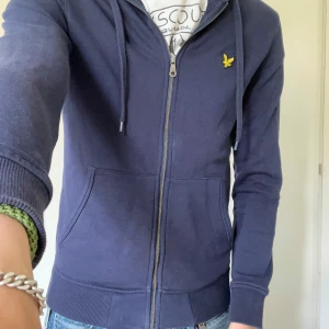 Mörkblå hoodie från Lyle & Scott - Säljer en mörkblå hoodie från Lyle & Scott med dragkedja och huva. Tröjan har två fickor framtill och den klassiska gula loggan på bröstet. Perfekt för en avslappnad stil och enkel att matcha med jeans eller joggers.
