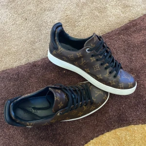 Louis Vuitton sneakers med monogram - Säljer ett par bruna Louis Vuitton sneakers med klassiskt monogrammönster och svarta skosnören. Skorna har vit platt sula och rund tå. Perfekta för dig som gillar lyxiga detaljer och streetstyle.