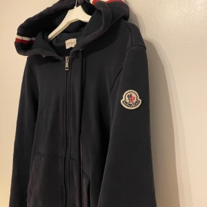 Mörkblå hoodie från Moncler. - Snygg mörkblå hoodie från Moncler med dragkedja framtill och fickor på sidorna. Jackan har huva med snörning och Moncler-logga på ärmen. Moncler märket är scannbart!  Pris är 1600kr ( är även på för byten)